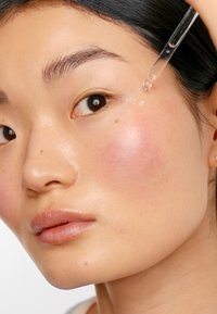 Un flacon compte-gouttes clair distribue un liquide sur la joue. La peau est lisse avec un éclat naturel ; une légère rougeur sur la joue et quelques taches de rousseur visibles.