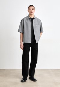 Cămașă cu mânecă scurtă, de culoare gri cu dungi, cu un buzunar, purtată peste un tricou negru cu guler rotund, asortată cu pantaloni și adidași negri.