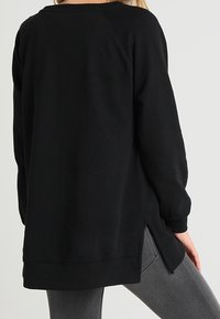 Persona che indossa un ampio top nero a maniche lunghe con fessure laterali, abbinato a jeans grigi aderenti, visto da dietro.