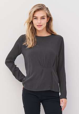 Blond kvinna med löst hår, iklädd en mörk långärmad topp och svarta byxor, som står mot en enfärgad vit vägg.