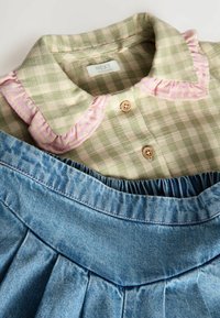 Chemise à carreaux vert clair avec un col froncé rose et des détails de boutons, superposée à une jupe en denim bleu avec un design plissé à la taille.