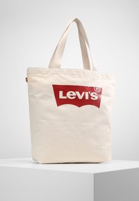 Beige canvas axelväska med röd Levi's-logotyp, två handtag upptill, fyrkantig form och slät yta. Inga ytterligare dekorationer eller mönster.