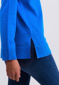 Helles blauer Pullover mit gerippten Bündchen und Saum, mit seitlichen Schlitzen für Bewegungsfreiheit. Die Textur wirkt weich und gestrickt.