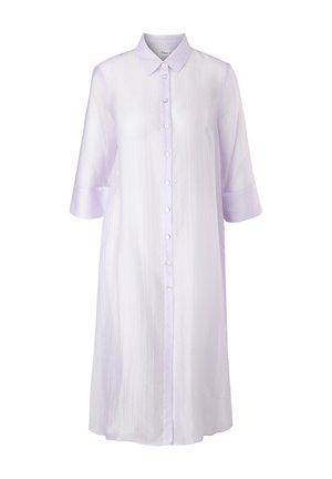 Femme portant une chemise longue lilas transparente boutonnée sur un pantalon blanc droit et des sandales noires à talons, debout devant un fond uni.