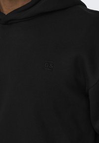 Hanorac negru cu glugă, fabricat dintr-un material moale. Prezintă un mic logo embosat "OS" pe piept. Design simplu și culoare uniformă.