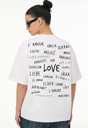Vrouw met donker haar in een knot, zwarte zonnebril en een wit T-shirt met het woord "love" in meerdere talen op de rug.