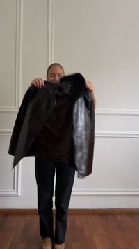 Personne debout à l'intérieur tenant une veste en cuir noire ouverte, portant un pantalon sombre et des chaussures marron clair devant un mur à panneaux blancs.