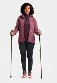 Giacca bordeaux con cappuccio sopra una maglia color bordeaux scuro, pantaloni neri e scarpe da ginnastica con accenti rossi. Tiene in mano bastoncini da trekking con punte verdi.