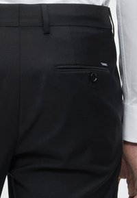 Κοντινό πλάνο ενός ατόμου που φοράει μαύρο παντελόνι με κουμπωτή πίσω τσέπη και φανερό μανίκι λευκού πουκάμισου με μακριά μανίκια.
