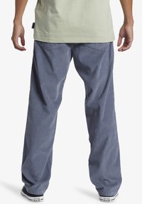 Blue corduroy pants with a loose fit, featuring a ribbed texture and wide legs. Worn with black sneakers showing branded details.