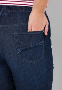 Jeans de mezclilla con un lavado oscuro y texturizado. Presentan un detalle de bolsillo rasgado y un corte relajado, con una mano visible descansando en el bolsillo trasero.