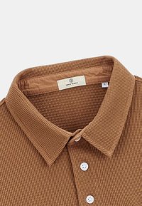 Polo shirt marron texturé avec un col, doté d'une fermeture à boutons et d'une étiquette à l'intérieur. Le tissu présente un design à côtes pour plus de détail.