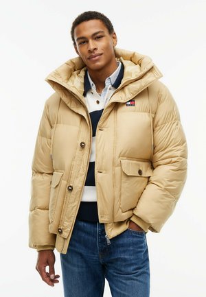 Joven con una chaqueta acolchada beige sobre una camisa de rayas y vaqueros azules, de pie con una mano en el bolsillo, sobre un fondo liso.