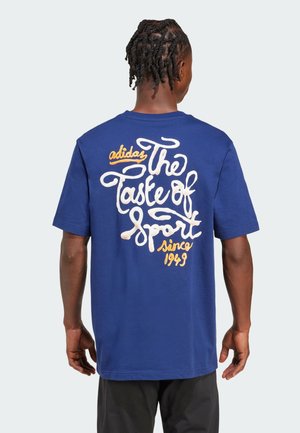 Blå bomulds T-shirt med hvid og orange kursiv tekst på ryggen. Designet inkluderer "Adidas" og "The Taste of Sport since 1949."