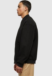 Chaqueta bomber negra con un ajuste relajado, puños acanalados y bolsillos laterales. La tela parece suave, con una textura lisa y un detalle mínimo.