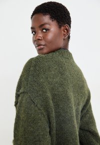 Groene gebreide trui met een gestructureerde afwerking, voorzien van een hoge halslijn en verlaagde schoudernaden, gezien van achteren.