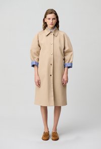 Cappotto beige con una vestibilità su misura, colletto grande, bottoni dorati e maniche a sbuffo. I polsini a righe blu e bianche aggiungono contrasto. Un paio di mocassini marroni.