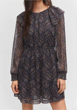 Frau trägt ein durchsichtiges, langärmliges, dunkel paisleygemustertes Kleid mit geraffter Taille und Manschetten, steht vor einem schlichten Hintergrund.