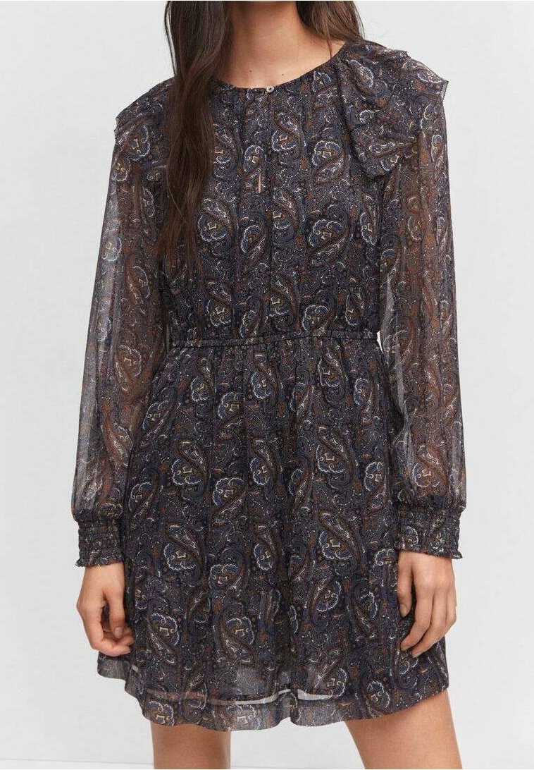 Femme portant une robe longue transparente à manches longues, de couleur foncée avec un motif cachemire, taille et poignets froncés, se tenant devant un fond uni.