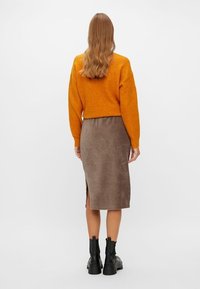 En orange kortärmad tröja och en brun kordfläkt midi kjol med en sidoklyfta, ihop med svarta ankelstövlar. Väggen är helt vit.