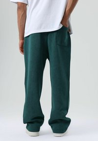 Loszittende groene sweatpants van zacht materiaal, met een achterzak en een afgeronde zoom. Gecombineerd met een wit T-shirt.