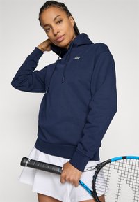 Marinblå hoodie i bomullsblandning, med en känguruficka, dragkedja på huvan och en liten grön logotyp på vänster bröst.