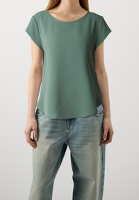 T-shirt a maniche corte in verde tenue, realizzato in tessuto morbido, con scollo rotondo e vestibilità rilassata, abbinato a jeans in denim blu chiaro.