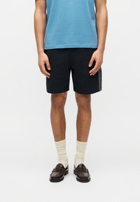 Homme portant un T-shirt bleu, un short noir, des chaussettes crème et des mocassins marron foncé, debout devant un fond blanc uni.
