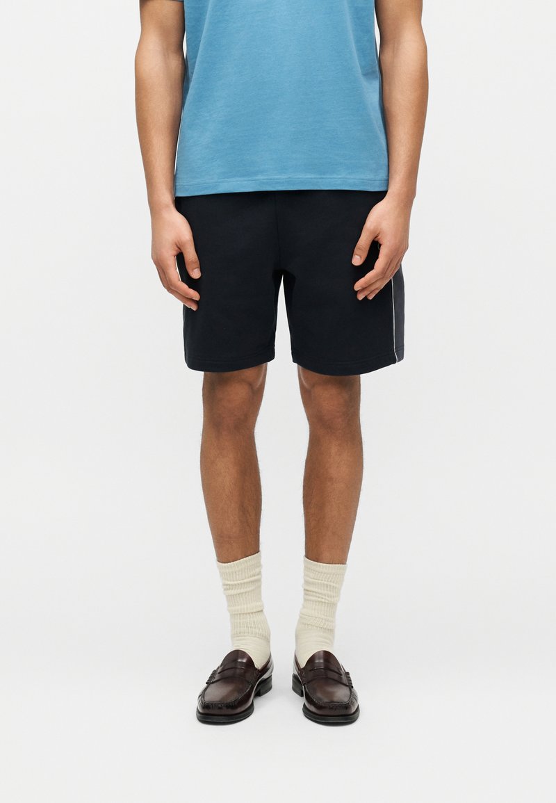 Homme portant un T-shirt bleu, un short noir, des chaussettes crème et des mocassins marron foncé, debout devant un fond blanc uni.