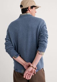 Pull en tricot bleu avec une finition texturée, doté de poignets et d'un ourlet côtelés. Superposé avec une chemise à carreaux en dessous. Fond neutre.