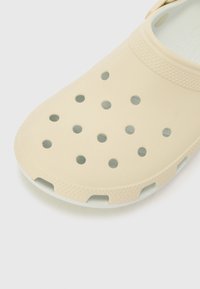 Lys beige kloss laget av fleksibelt materiale; har runde ventilasjonshull og en teksturert såle for grep. Jevn overflate generelt.