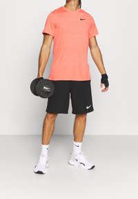 Camiseta de manga corta en coral jaspeado de Nike; pantalones cortos negros con logo; sosteniendo mancuernas negras; calcetines blancos y zapatillas con acentos de marca.