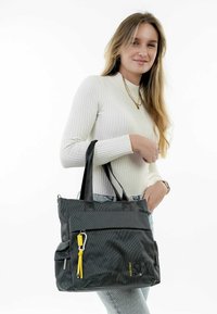 Black textured tote bag with a zippered top, front pocket, yellow accent, and black straps. Hardware includes a silver clip and zipper pull.