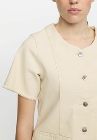 Beige klänning i bomullsblandning med korta ärmar, fransad fåll och sex metallknappar. Har en rund halsringning och två framfickor.