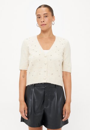 Crème korte mouwen cardigan met V-hals, voorzien van textuurdetails in kralen en vijf knopen aan de voorkant, gecombineerd met zwarte geplooide shorts.