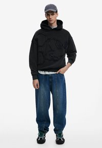 Sort sweatshirt med præget Mickey Mouse design, parret med løse blå denimjeans. Accessories inkluderer en grå kasket og sorte sneakers.