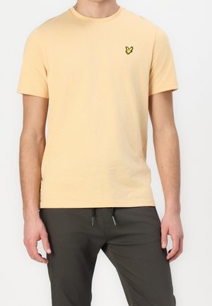 Homme portant un t-shirt jaune pâle à manches courtes avec un petit logo d'oiseau jaune sur la poitrine gauche, associé à un pantalon foncé.