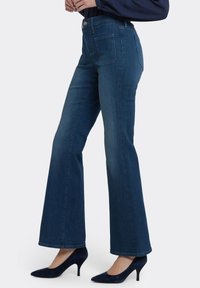 Mörkblå utställda jeans med en slät denimtextur, hög midja och två bakfickor. Kombinerade med marinblå högklackade skor.