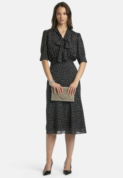 Črna obleka z belimi pikami, z ovratnikom z mašno, kratkimi rokavi in razširjenim krilom. Kombinirana z metalik torbico in črnimi petami.