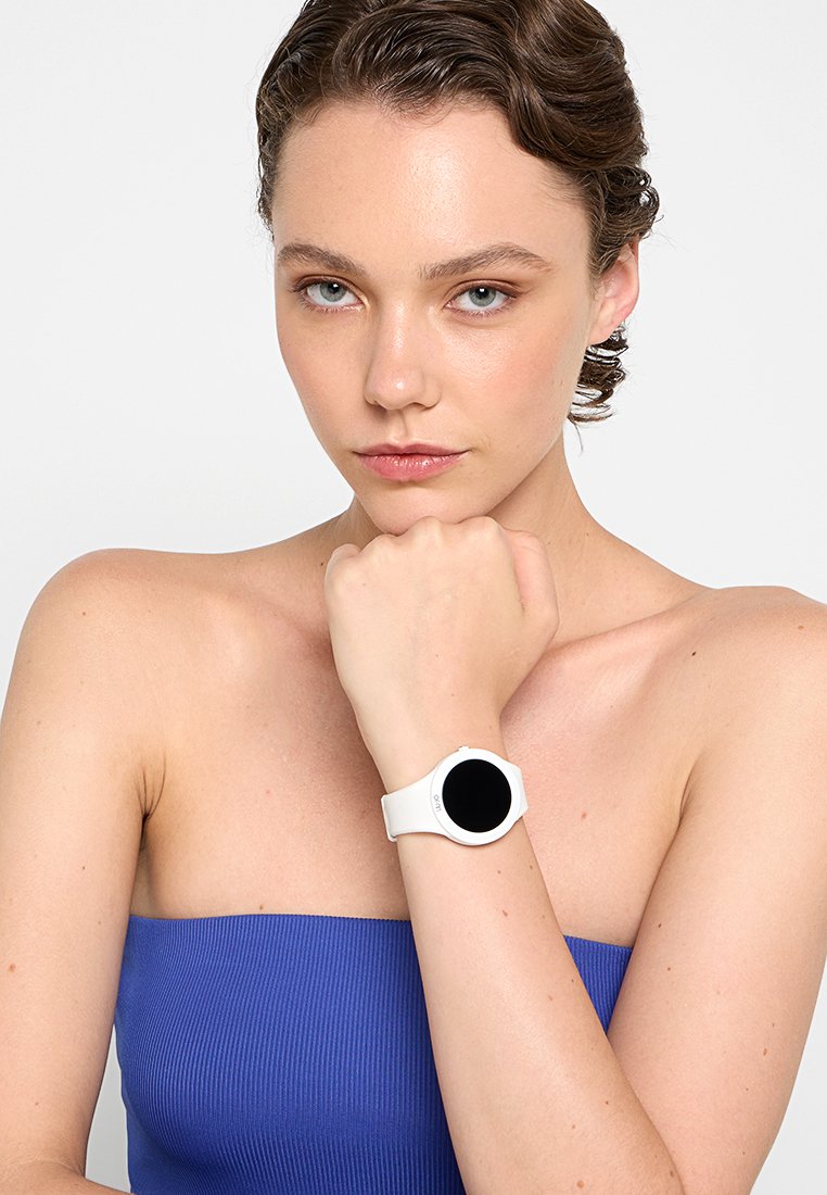 Montre connectée ronde avec un bracelet en silicone blanc, dotée d'un écran noir. L'utilisateur porte un haut bleu à côtes, le bras reposant dans une position neutre.