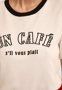 Kremowa koszulka z czarną ściągaczem na dekolcie. Posiada wyszywany tekst: "UN CAFÉ s'il vous plaît" w kontrastującym czarnym haftem.