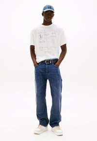 Camiseta gráfica blanca con el texto de Tommy Jeans, pantalones cargo de denim azul, cinturón negro con acentos rojos y azules, y zapatillas beige. Gorra de denim.