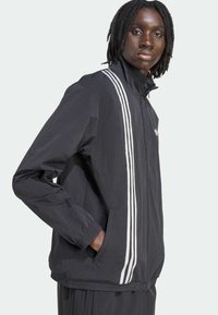 Black jacket with white three-stripe design running down sleeves, zip front, and elastic cuffs. Lightweight fabric with a relaxed fit.