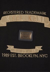 Texte brodé « Registered Trademark KARL KANI », patch logo métal et « 5-23-68 1989 Est. Brooklyn, NYC » sur tissu noir.