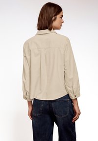 Femme debout, de dos, portant une chemise beige à manches longues et un jean bleu foncé sur un fond blanc uni.