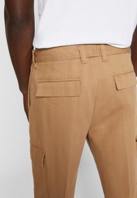 Khaki Cargo-Hosen mit geradem Schnitt, zwei hinteren Klappentaschen und subtilen Stickdetails. Hergestellt aus leichtem, strukturiertem Stoff.
