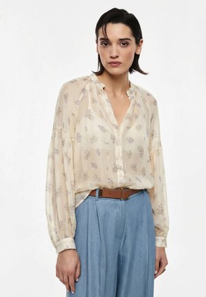 Donna con una camicetta crema trasparente con motivo floreale e maniche a palloncino, infilata in pantaloni blu a vita alta con una cintura marrone.