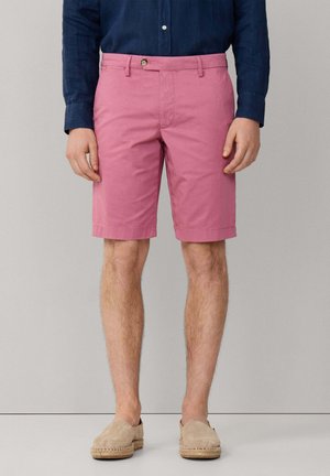 Hombre con pantalones cortos rosas hasta la rodilla, camisa de manga larga azul marino y alpargatas beige, de pie contra un fondo liso.