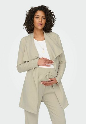 Veste beige à superposition avec un avant ouvert, associée à un top blanc et un pantalon smocké assorti. Texture lisse, design minimaliste.