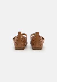 Zapatos de cuero marrón claro con detalles perforados, un tacón bajo y un cierre de hebilla decorativa en el lateral. Parte trasera redondeada y textura suave.